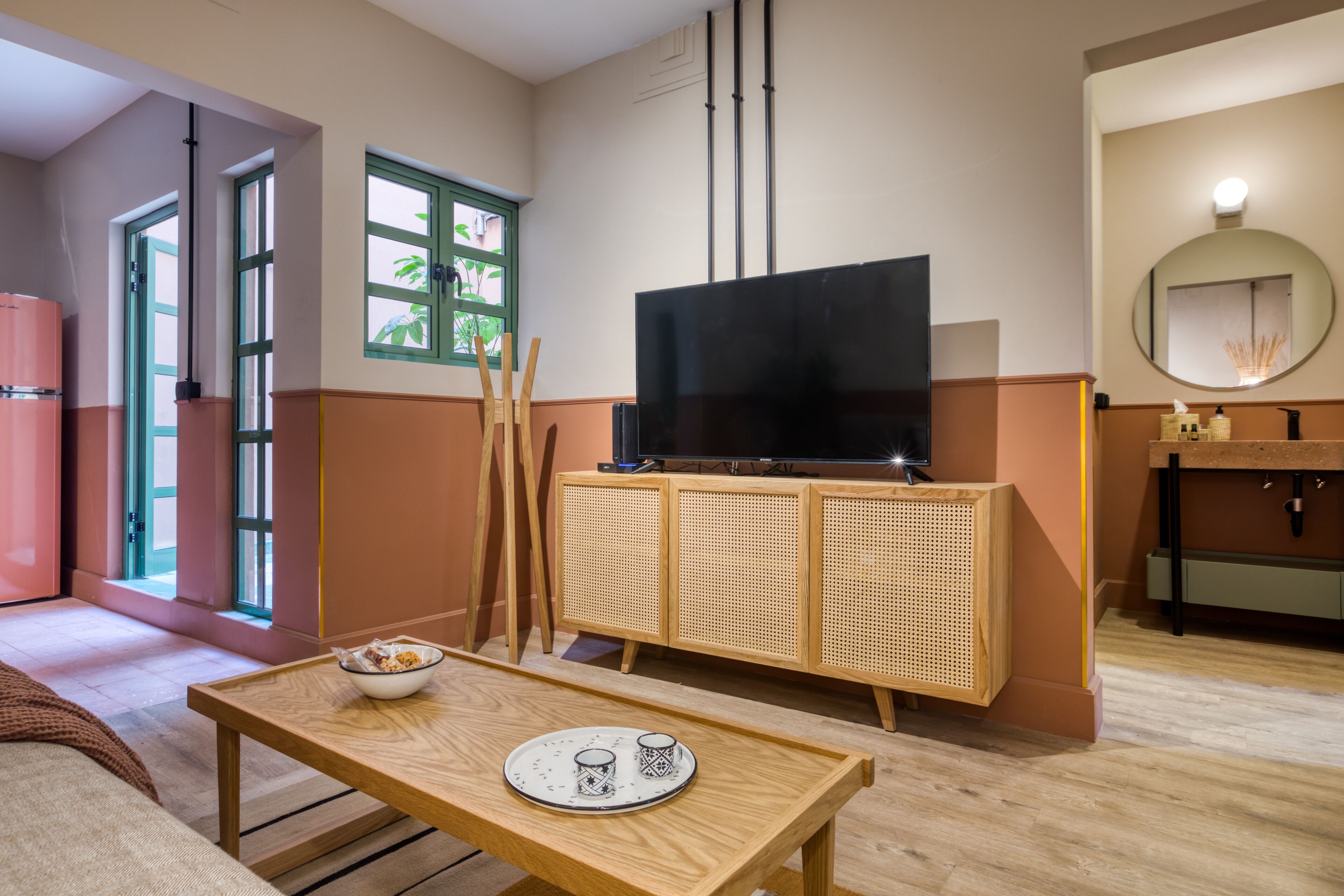 A modern hotel room with a wooden table, a television on a rattan cabinet, a mirror, and a window with a view of greenery.