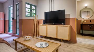 Una habitación de hotel moderna con una mesa de madera, un televisor sobre un mueble de ratán, un espejo y una ventana con vistas a la vegetación.