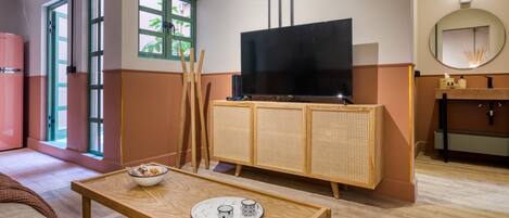 A modern hotel room with a wooden table, a television on a rattan cabinet, a mirror, and a window with a view of greenery.