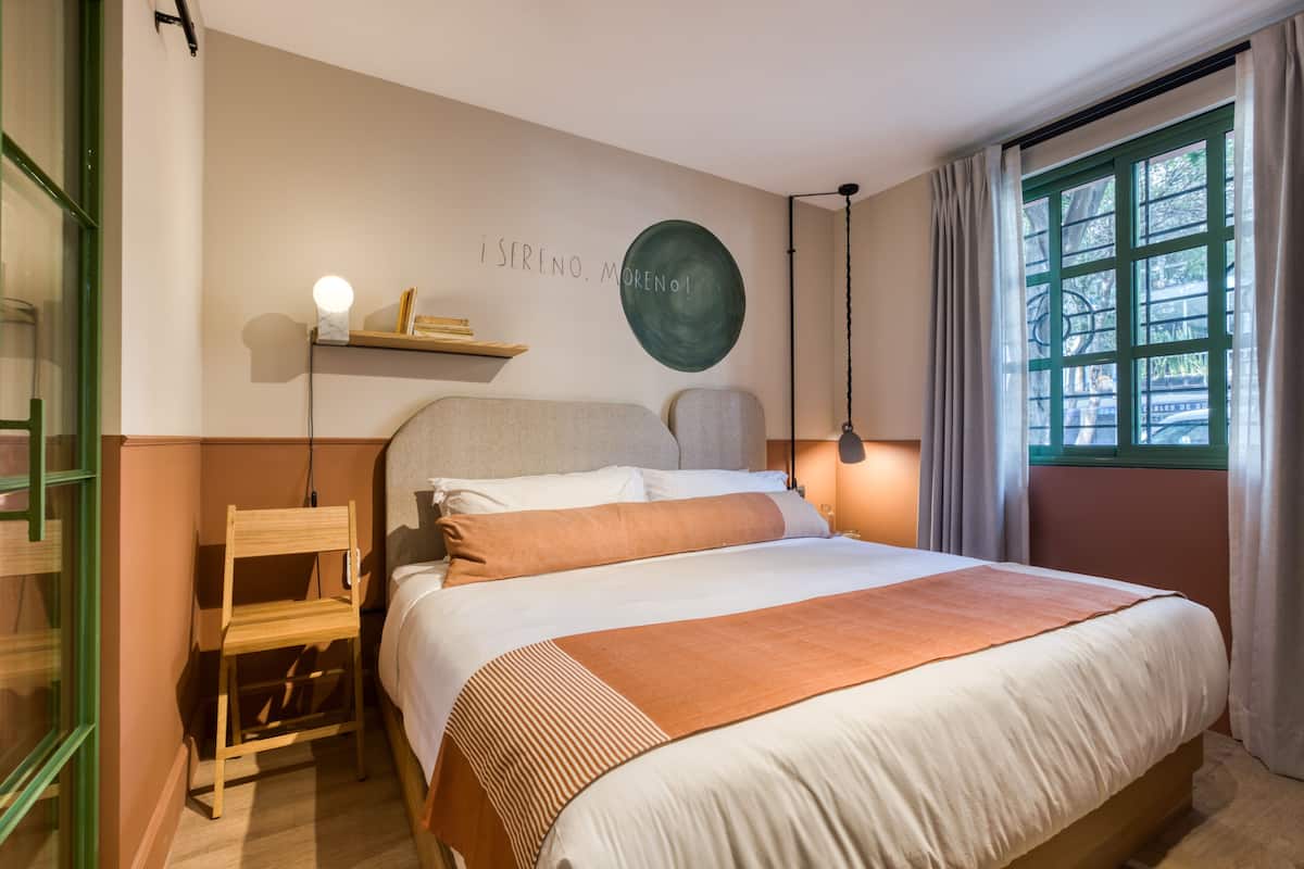 A neatly arranged bedroom with a bed, bedside table, lamp, and a wall-mounted shelf with a book and a light.
