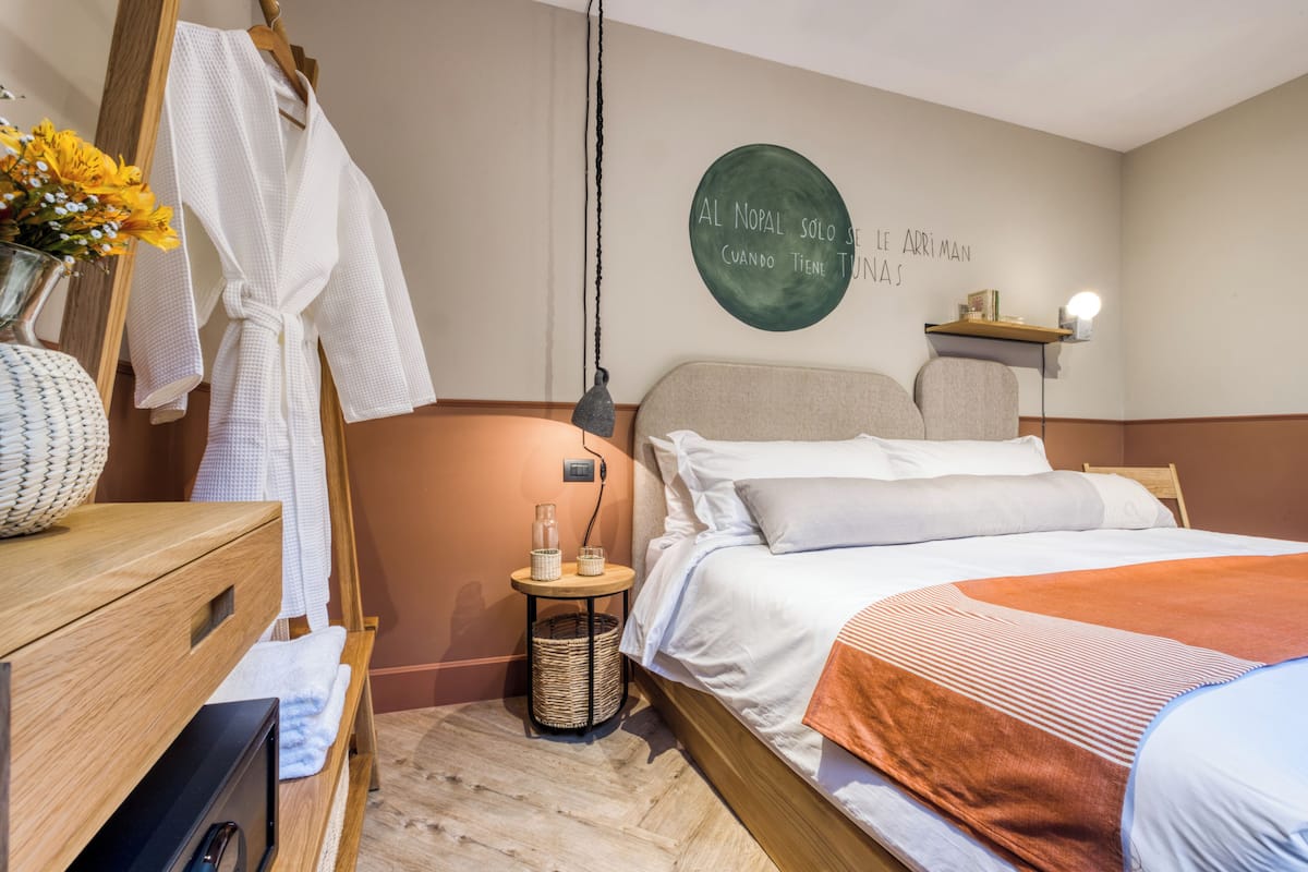 A hotel room with a bed, a wooden dresser, a robe hanging on a rack, a wall with a circular sign, and a small table with a lamp.