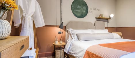 Una habitación de hotel con una cama, un aparador de madera, una bata colgada en un perchero, una pared con un letrero circular y una mesita con una lámpara.