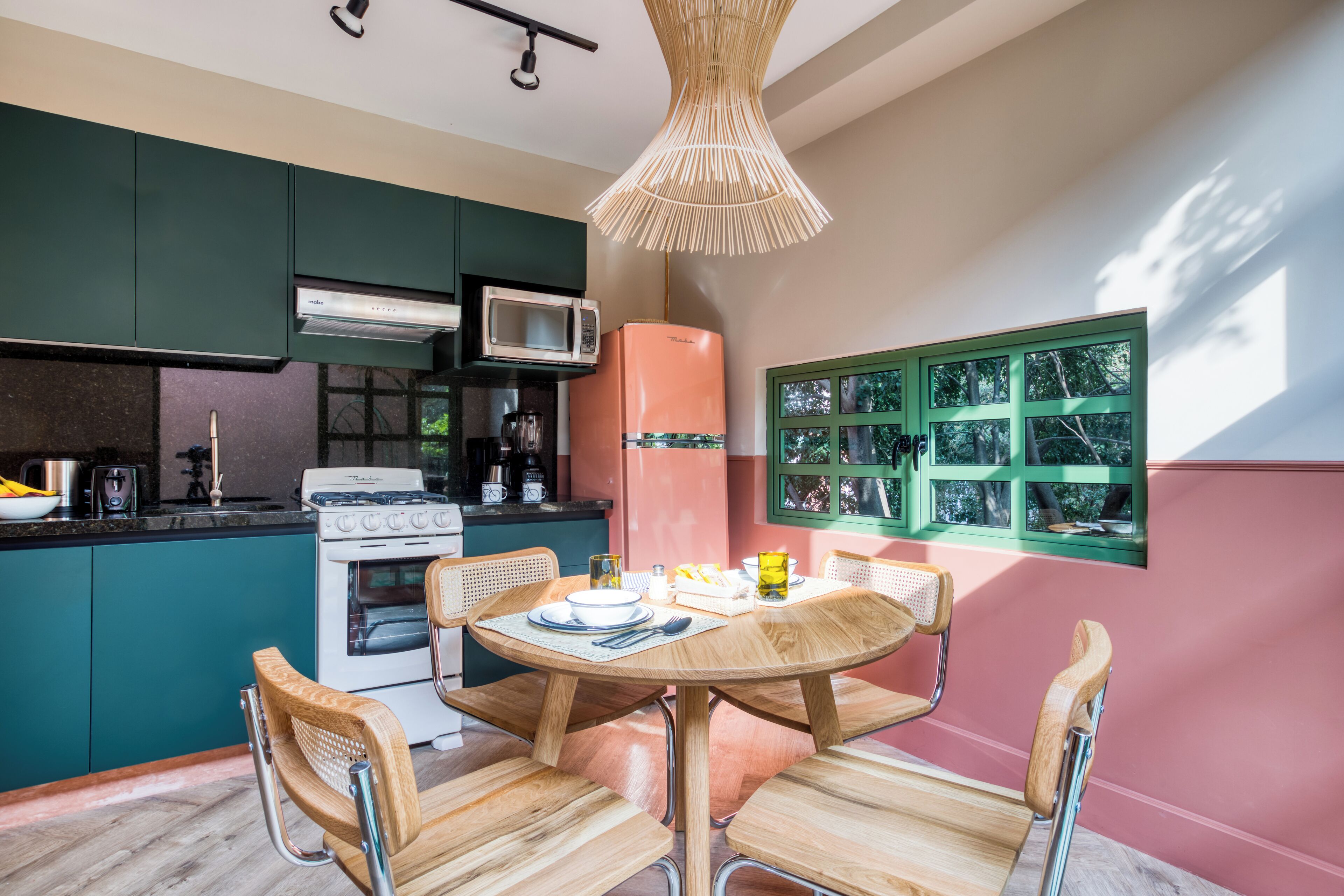 Una cocina con armarios azul verdoso, un refrigerador rosa y una mesa de comedor con sillas de madera.