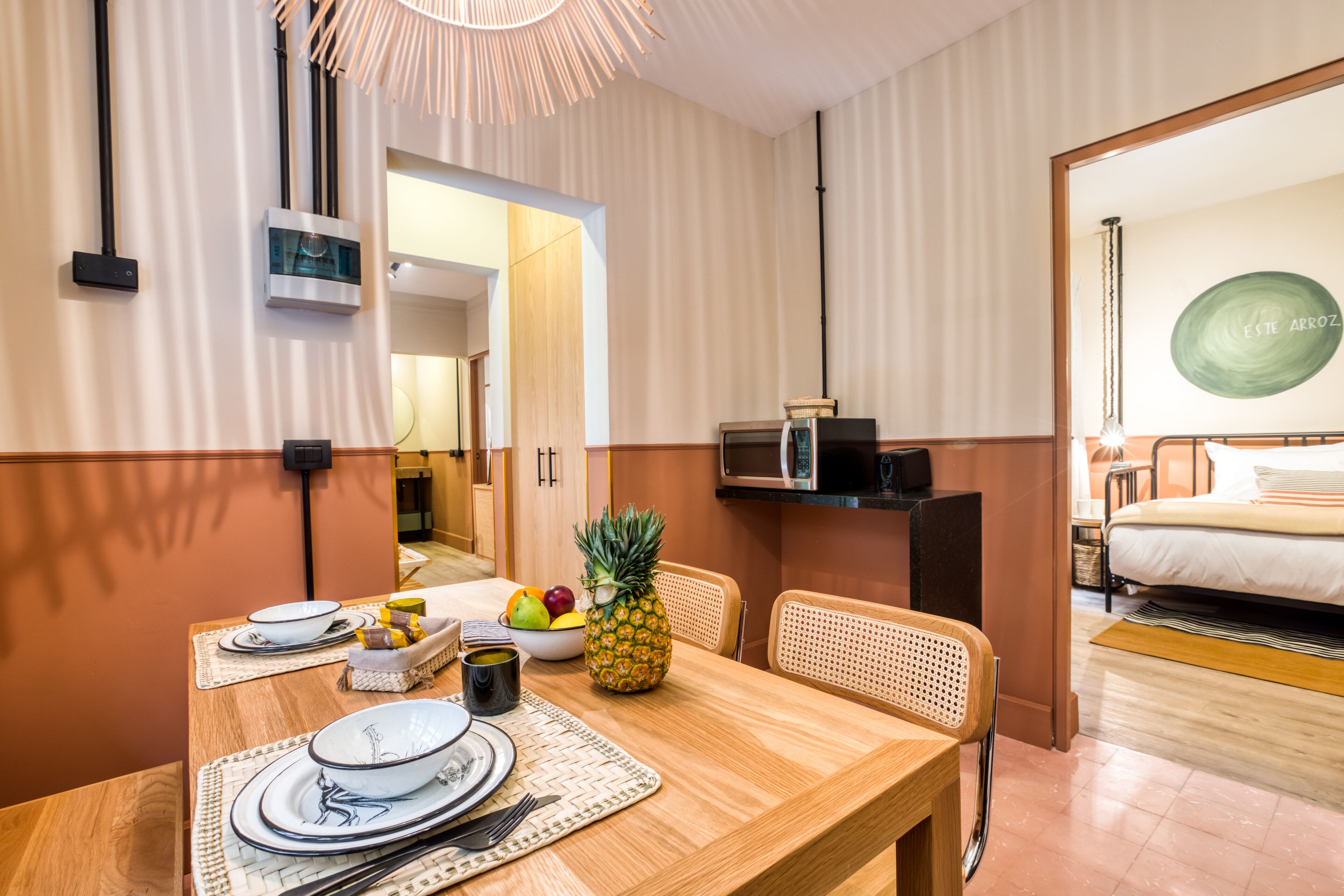 Ein Hotelzimmer mit einem Holztisch, gedeckt mit Tellern, Schüsseln und einer Ananas. Es gibt eine Mikrowelle, einen kleinen Fernseher und ein Bett mit einer grünen, runden Wanddekoration.