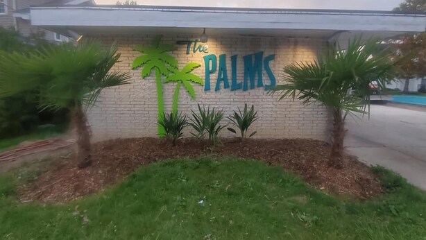 A building with a sign that reads "The PALMS" on a brick wall, surrounded by palm trees and grass. - The Palms Motel (Geneva)