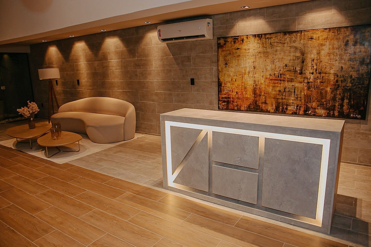 Una zona de recepción moderna con diseño minimalista, que incluye un mostrador de recepción, un sofá, una pequeña mesa con lámpara y un cuadro abstracto de gran tamaño en la pared.