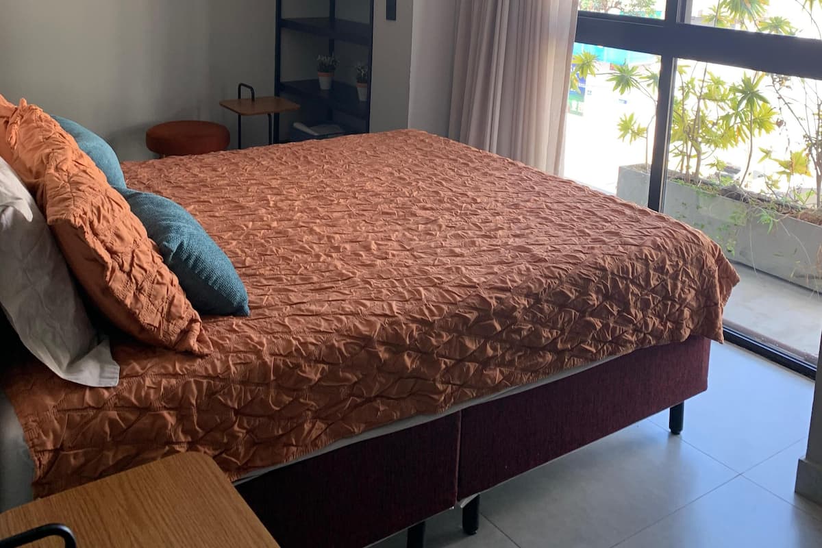 Un dormitorio con una cama grande, una mesita de noche, una silla, un armario empotrado y un ventanal con vistas a edificios y árboles.
