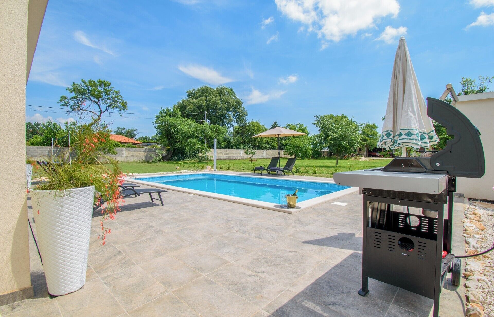 A swimming pool with lounge chairs and a barbecue grill in a backyard.