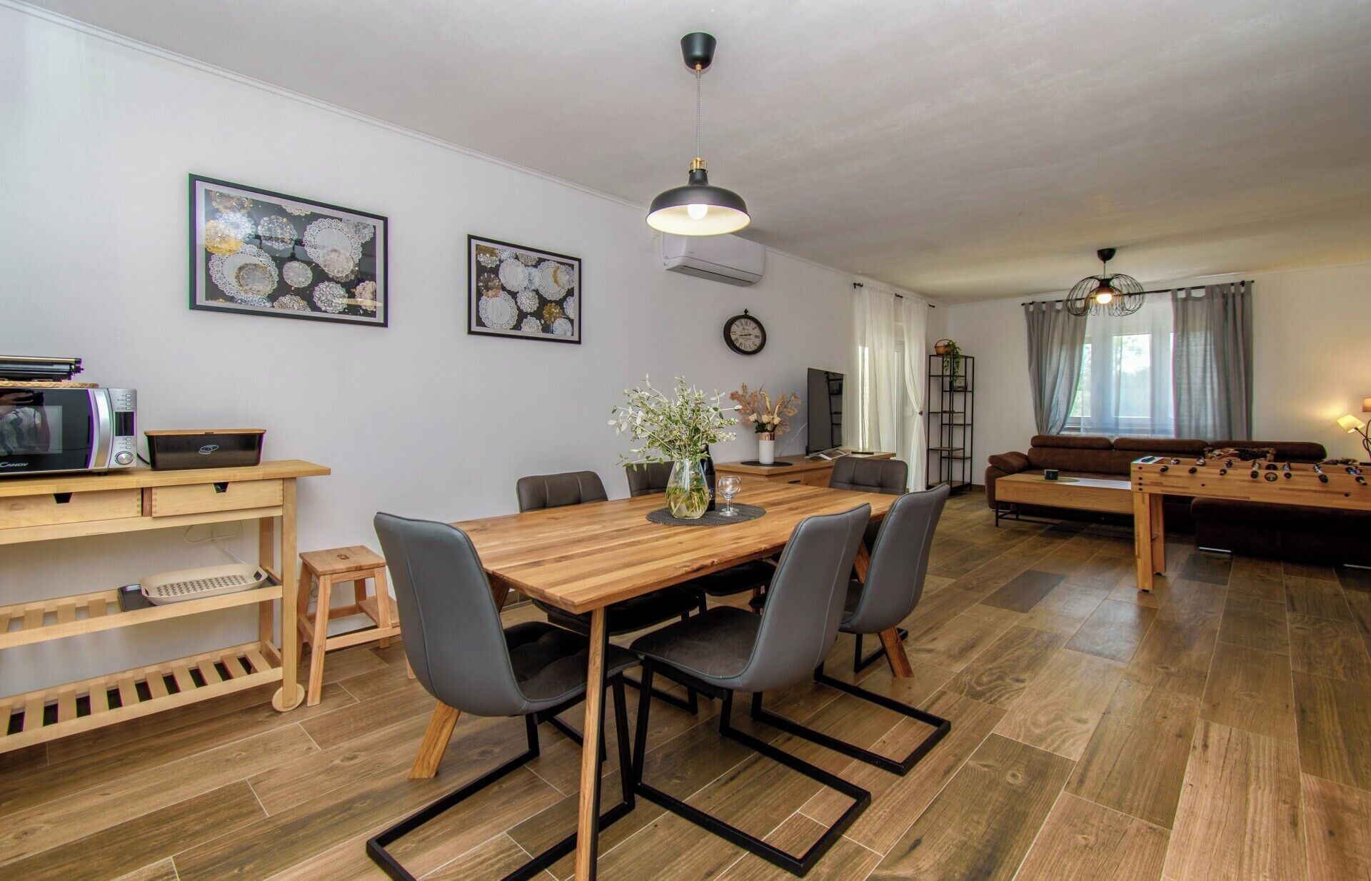 Ein modernes Wohnzimmer mit einem Holzesstisch, grauen Stühlen, einer Couch und einem Tischkicker. An der Wand hängen zwei gerahmte Kunstwerke, eine Uhr und eine Deckenleuchte.