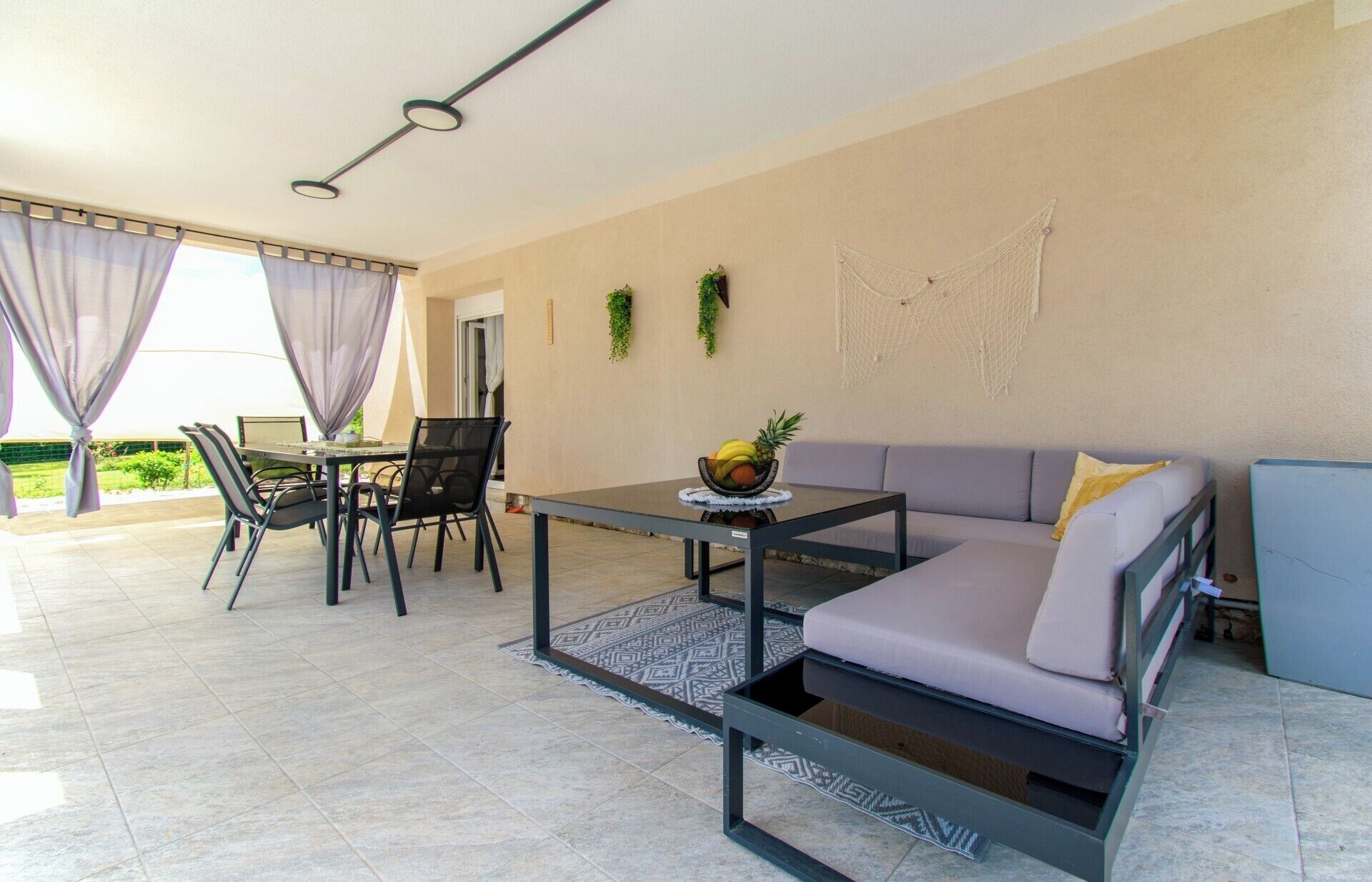 Una sala de estar moderna con un sofá, una mesa de comedor y sillas. Hay una mesita con un frutero encima. La habitación tiene un ventanal con cortinas, una planta colgante en la pared y un tapiz de macramé.
