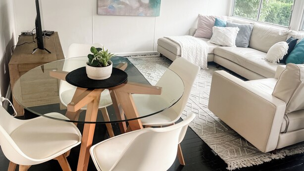 A modern living room with a glass dining table, white chairs, a beige sofa, and a large abstract painting on the wall.