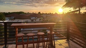 A terrace with a wooden table and stools, overlooking a residential area and palm trees during sunset.