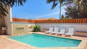 A rectangular swimming pool with clear blue water, surrounded by a stone patio. There are four white lounge chairs aligned along the pool's edge.