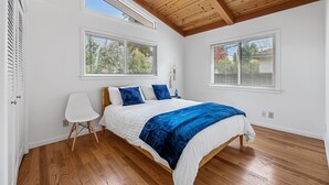 A bedroom with a wooden ceiling, two windows with blinds, a bed with a blue and white bedspread, a white chair, and wooden flooring.