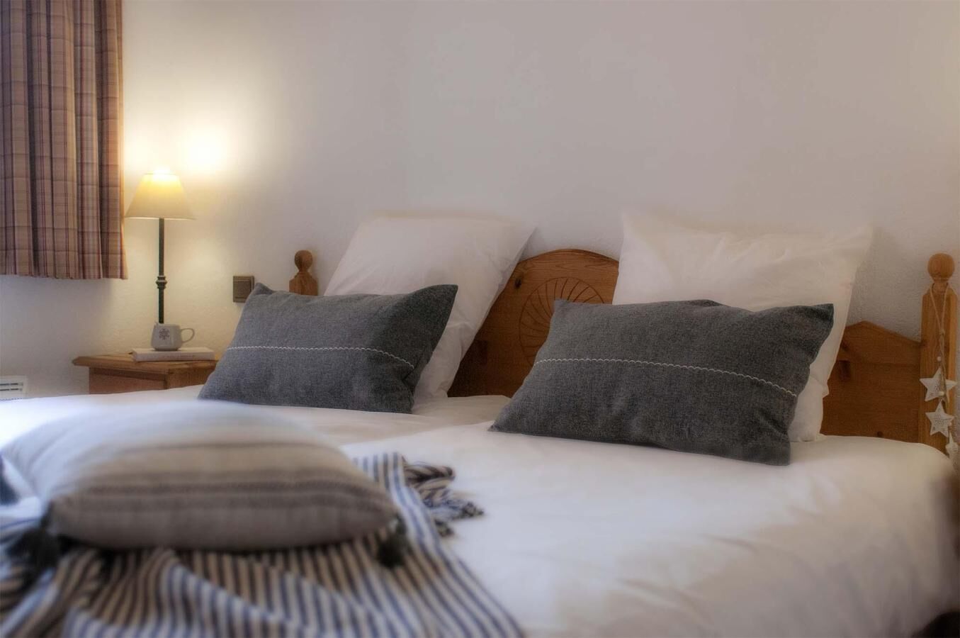 Een keurig opgemaakt bed met twee kussens, een nachtkastje met een lamp en een raam met gordijnen.