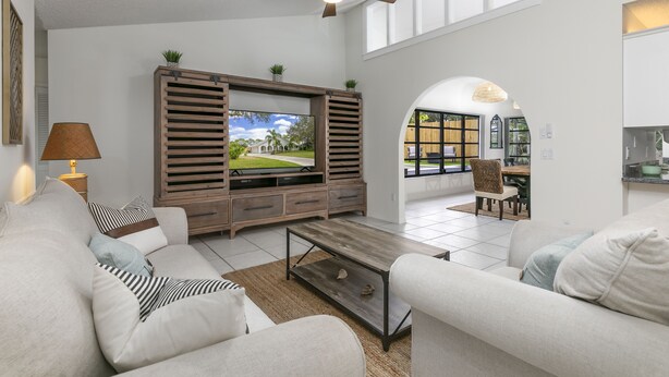 A spacious living room with a large flat-screen TV, a ceiling fan, and a dining area visible through an archway. - Peaceful and Spacious Pool home in Jensen Beach! RV-Boat Prkg-Fenced Yrd (Jensen Beach)