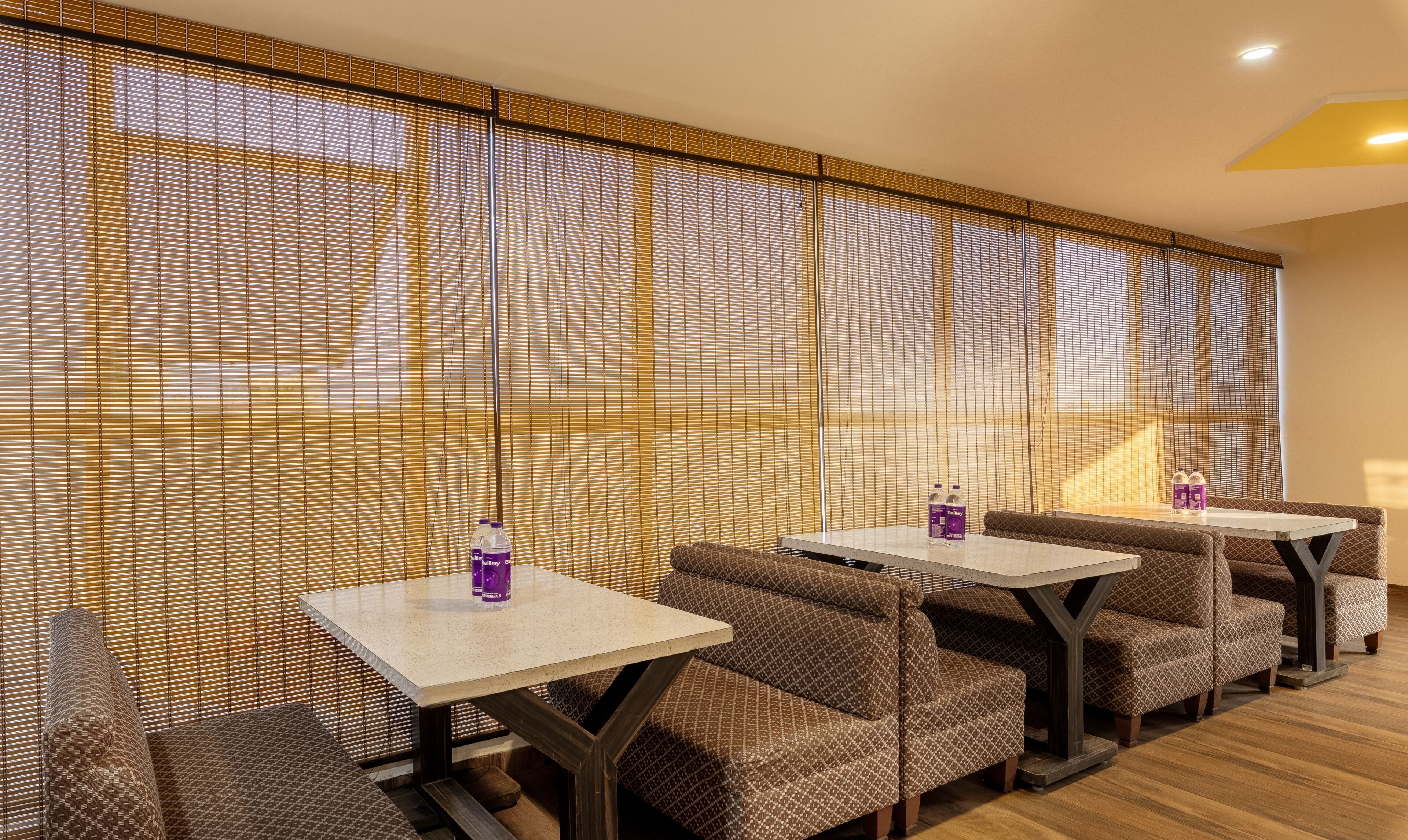 A dining area with tables and cushioned seating, a wooden floor, and a wall with vertical blinds.