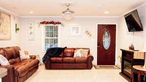 A living room with two brown leather sofas, a chandelier, a wooden door with a glass panel, and a television mounted on the wall. - Cozy Country Retreat Ranch Style Home, private hot tub, pet friendly (Sanford)