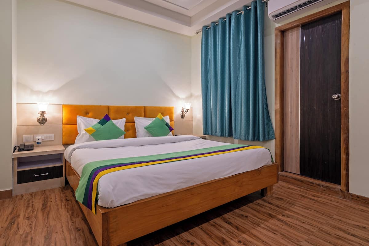 Een net opgemaakt bed met een houten hoofdeinde, een nachtkastje met een lamp en een groot raam met gordijnen.
