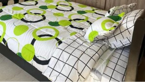 Une chambre à coucher avec un lit recouvert d’une couvre-lit à motifs géométriques, une armoire en bois dotée de plusieurs compartiments et une porte sobre en arrière-plan.