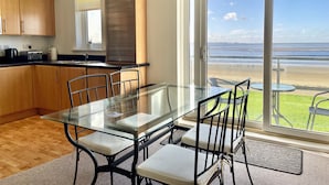 A modern kitchen with wooden cabinets, a glass dining table, and a view of the beach through large sliding glass doors. - Modern Beachfront Bliss with Spectacular Views (Llanelli)