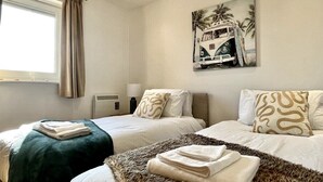 A hotel room with two beds, beige curtains, a painting of a vehicle on the wall, and a window with a view of palm trees. - Modern Beachfront Bliss with Spectacular Views (Llanelli)