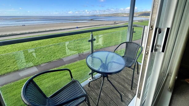 A balcony with a glass table and chairs, overlooking a beach and grassy area. - Modern Beachfront Bliss with Spectacular Views (Llanelli)