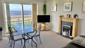 A living room with a glass table and chairs, a flat-screen TV, a fireplace, and a view of the ocean. - Modern Beachfront Bliss with Spectacular Views (Llanelli)