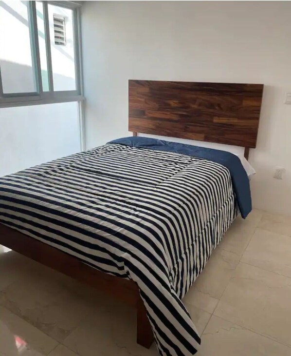Una cama con colcha a rayas y cabecera de madera en una habitación con paredes blancas y una ventana.