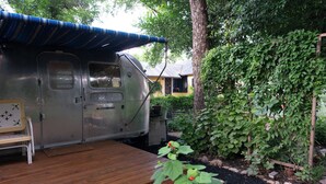 A stationary RV with a covered porch, a bench, and a garden area with plants and a tree.