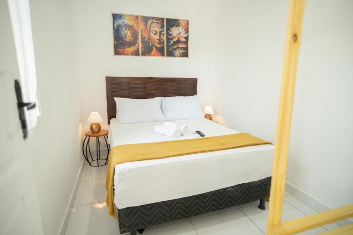 Un lit bien fait, avec une tête de lit en bois, du linge de lit blanc et un plaid jaune. Deux lampes de chevet sont placées de chaque côté du lit.