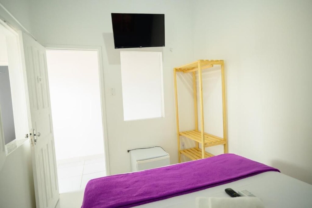 Une chambre avec un lit, un téléviseur fixé au mur, une armoire en bois et une petite table blanche.