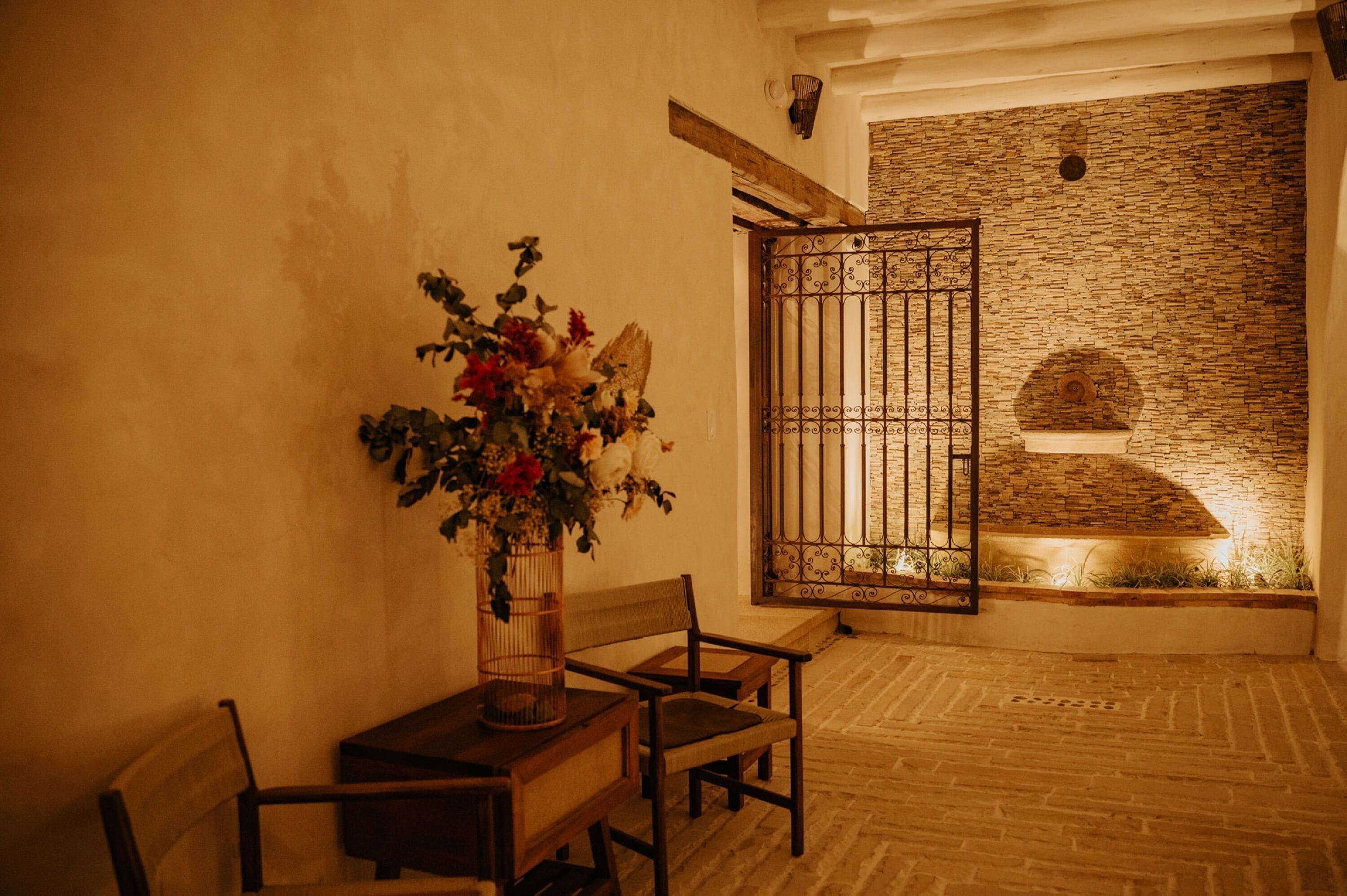 Una habitación con piso de madera, un banco de madera, una mesa con un florero y una pared de piedra con una reja de metal.