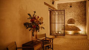 Una habitación con piso de madera, un banco de madera, una mesa con un florero y una pared de piedra con una reja de metal.