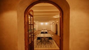 Una habitación larga y estrecha con techo alto y vigas de madera, que conduce a un comedor con mesas y sillas.