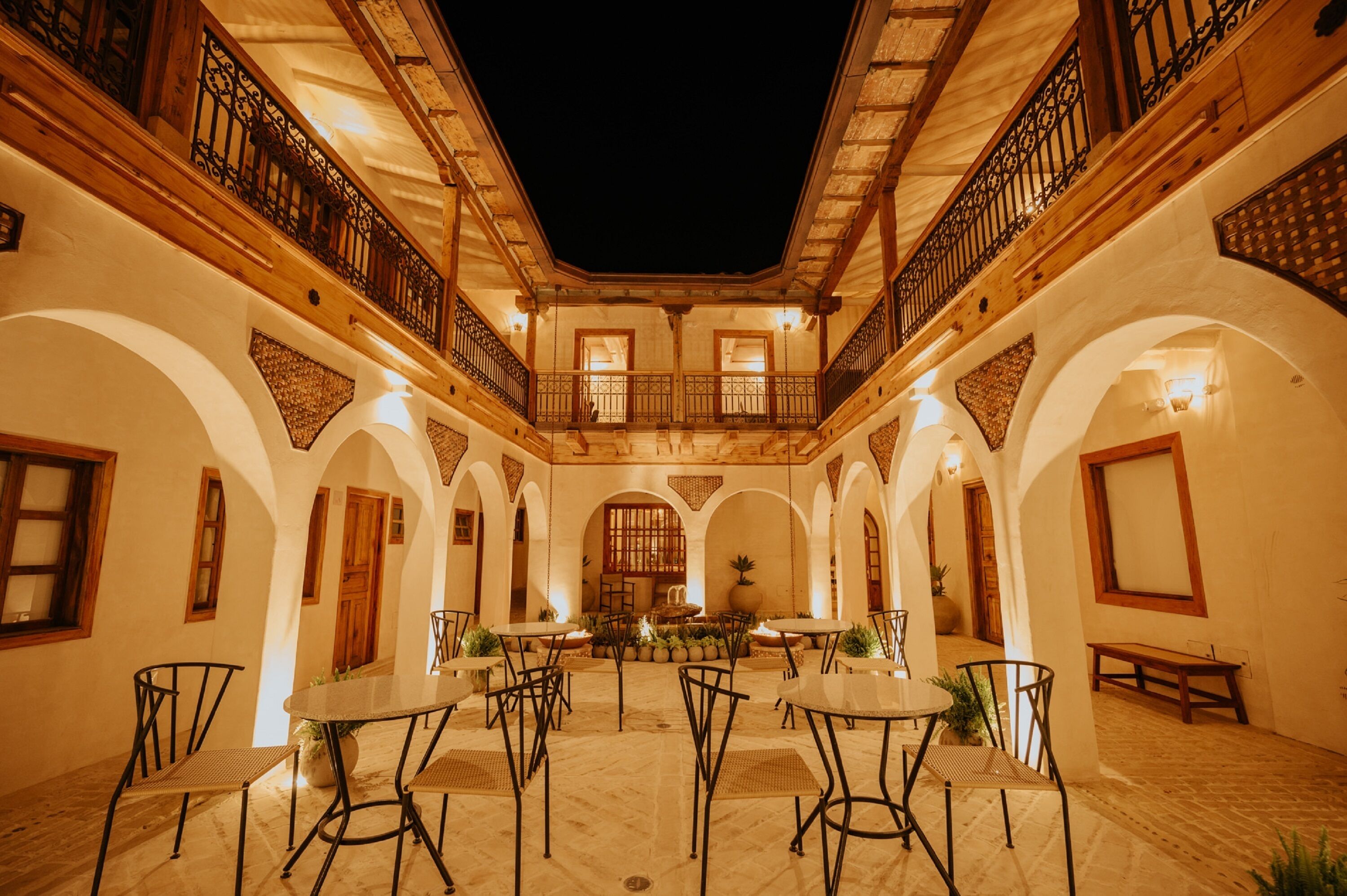 An indoor courtyard with a central fountain, surrounded by tables and chairs, and wooden balconies with wrought iron railings.
