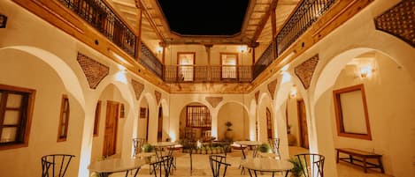 An indoor courtyard with a central fountain, surrounded by tables and chairs, and wooden balconies with wrought iron railings.