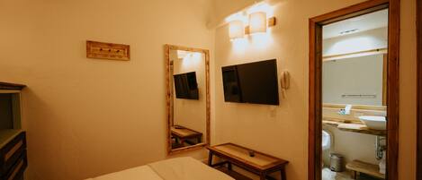 A hotel room with a bed, a small wooden bench, a TV, a mirror, and a bathroom visible through an open door.