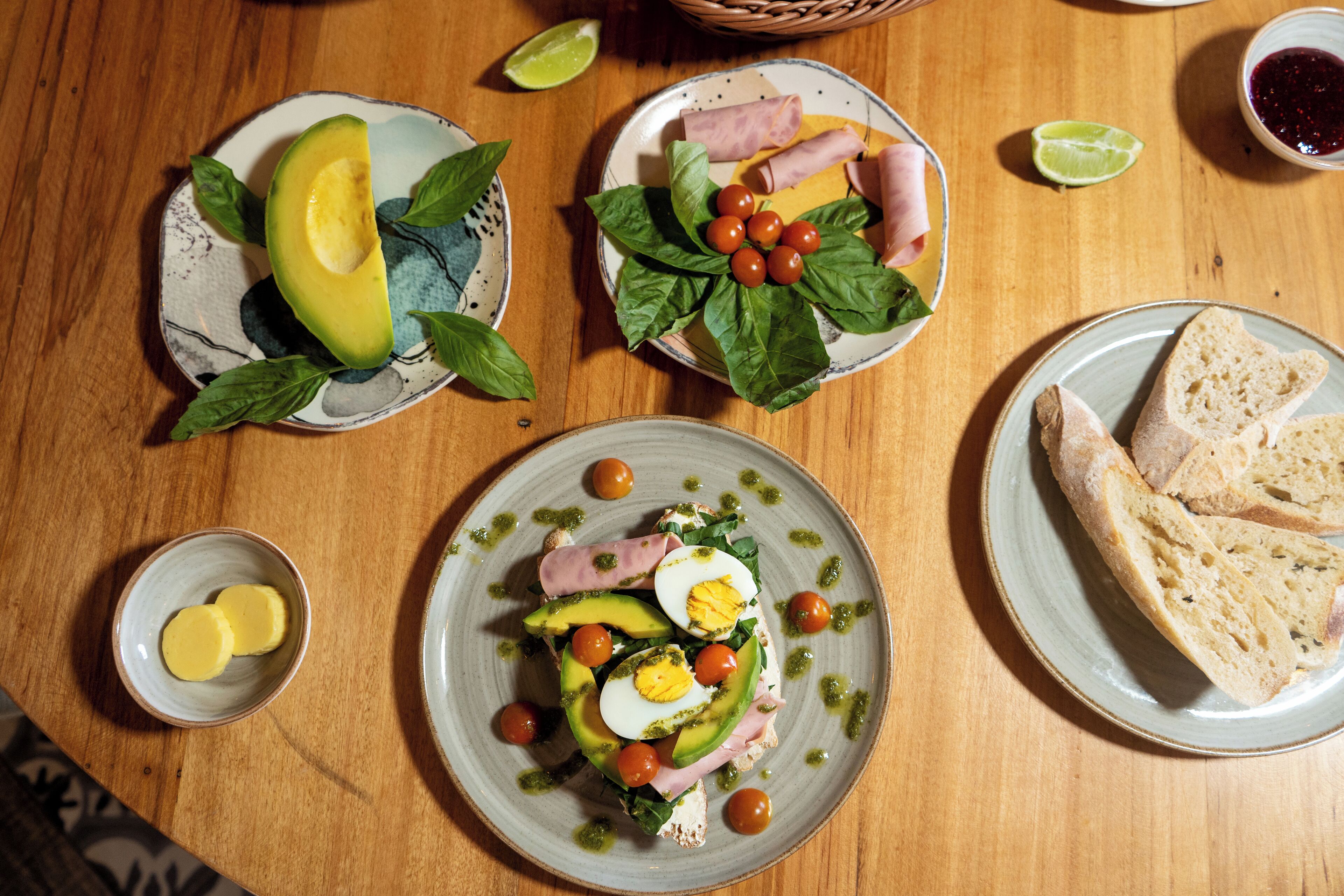 A wooden table with various plates of food, including avocado, eggs, tomatoes, and bread.