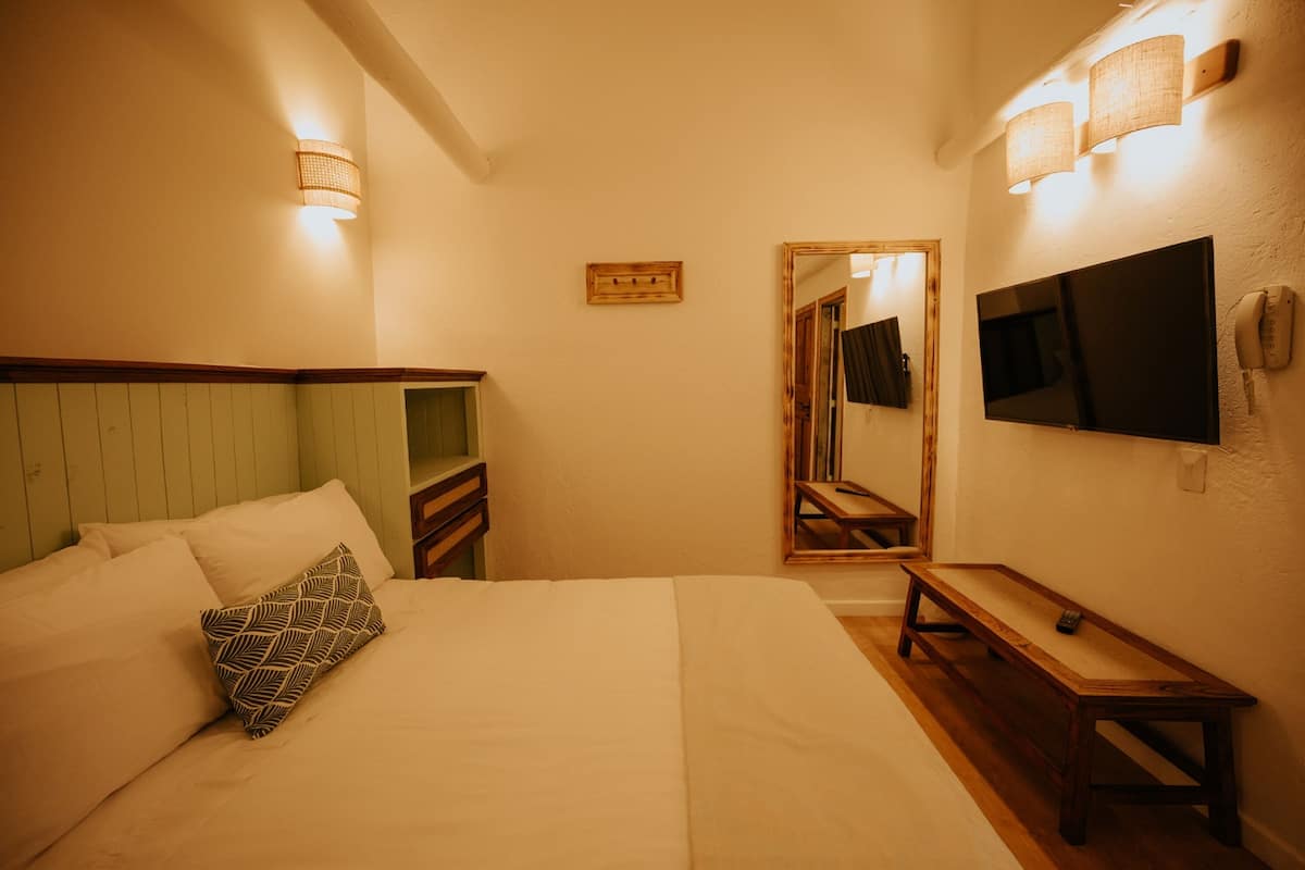 A hotel room with a bed, a wooden bench, a TV mounted on the wall, and a mirror.