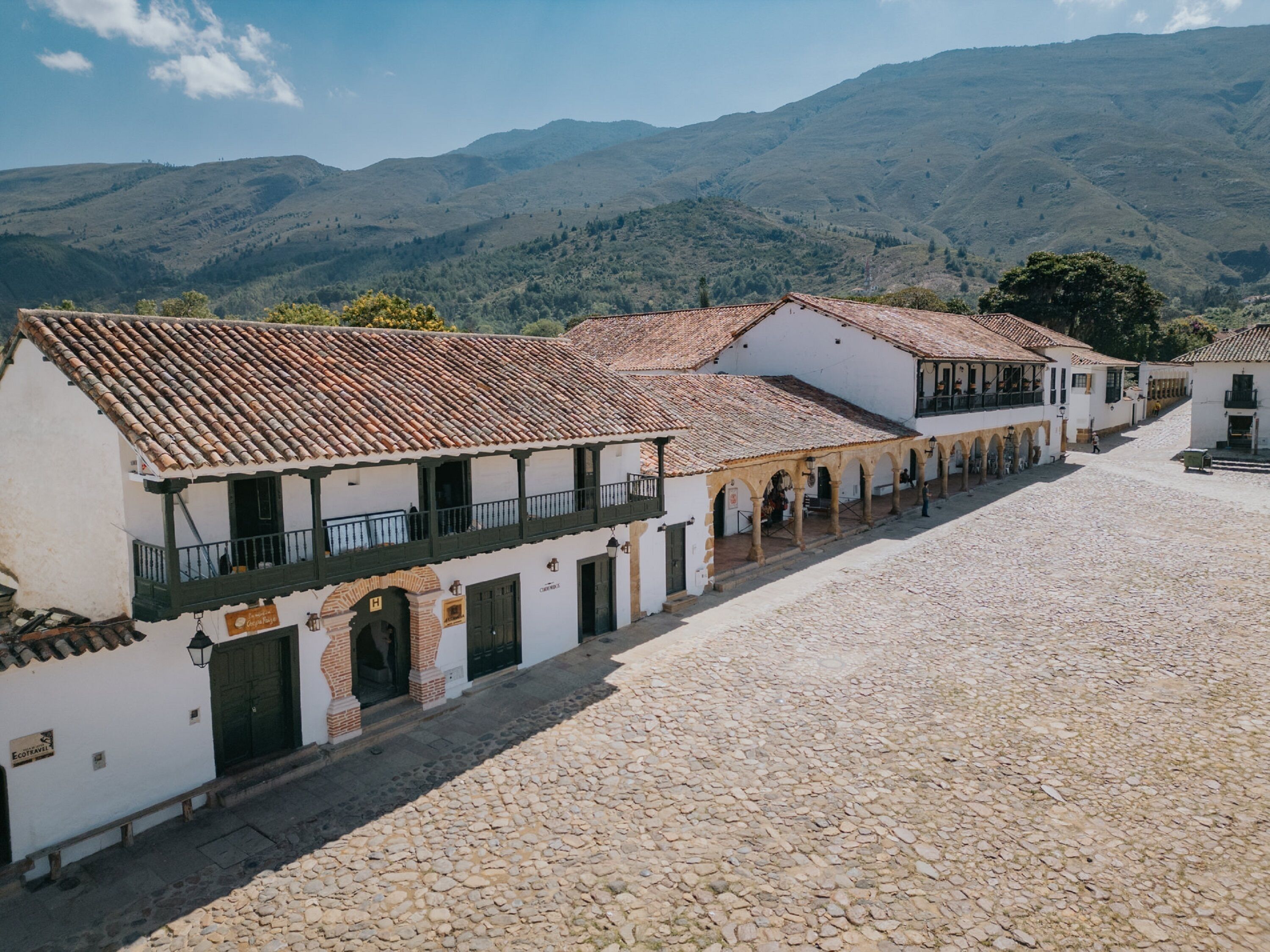 A cobblestone street lined with traditional buildings featuring tiled roofs and white walls, set against a backdrop of green hills.