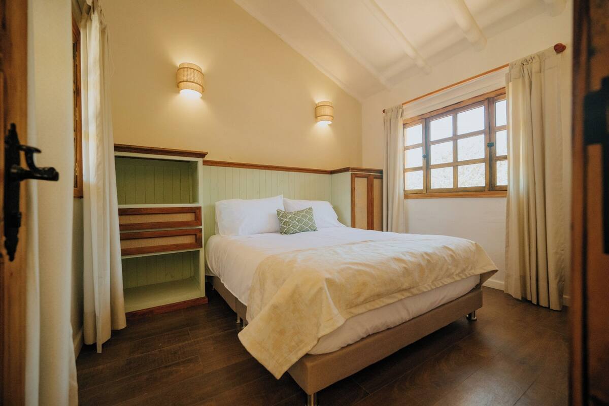A bedroom with a bed, wooden headboard, two wall sconces, a window with curtains, and a green upholstered bench.