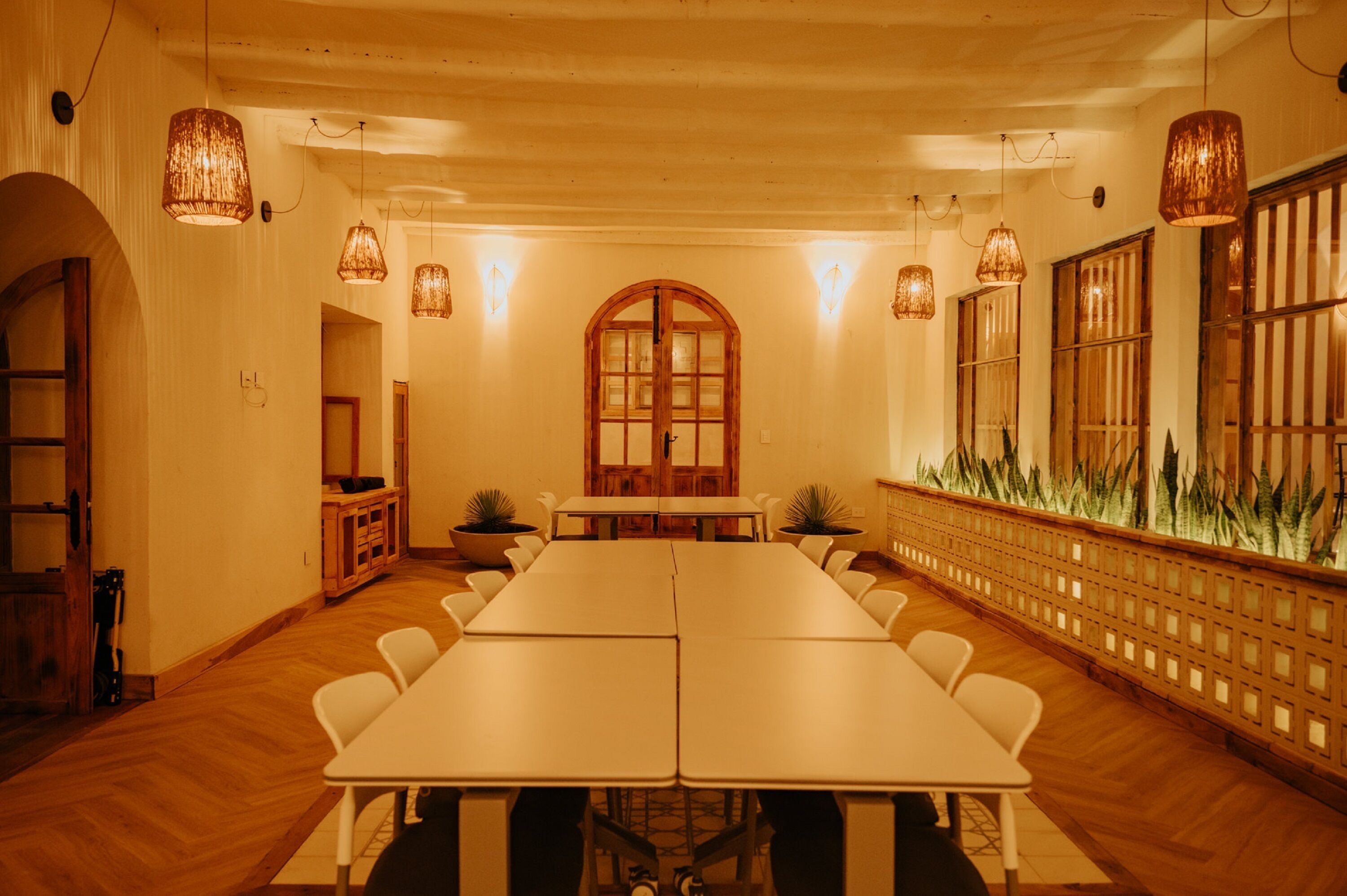 A conference room with a long table, white chairs, hanging lamps, and a wooden cabinet.