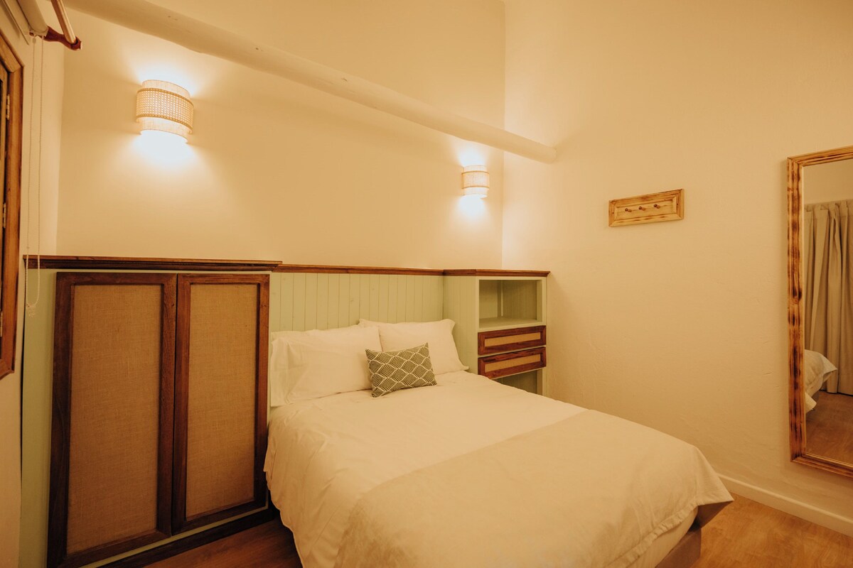 Un dormitorio con cama, mesitas de noche, un espejo e iluminación en la pared.