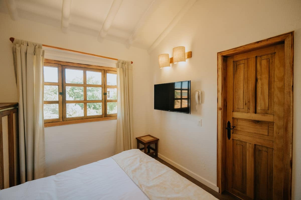 A bedroom with a bed, a window with curtains, a wooden door, and a mounted TV.