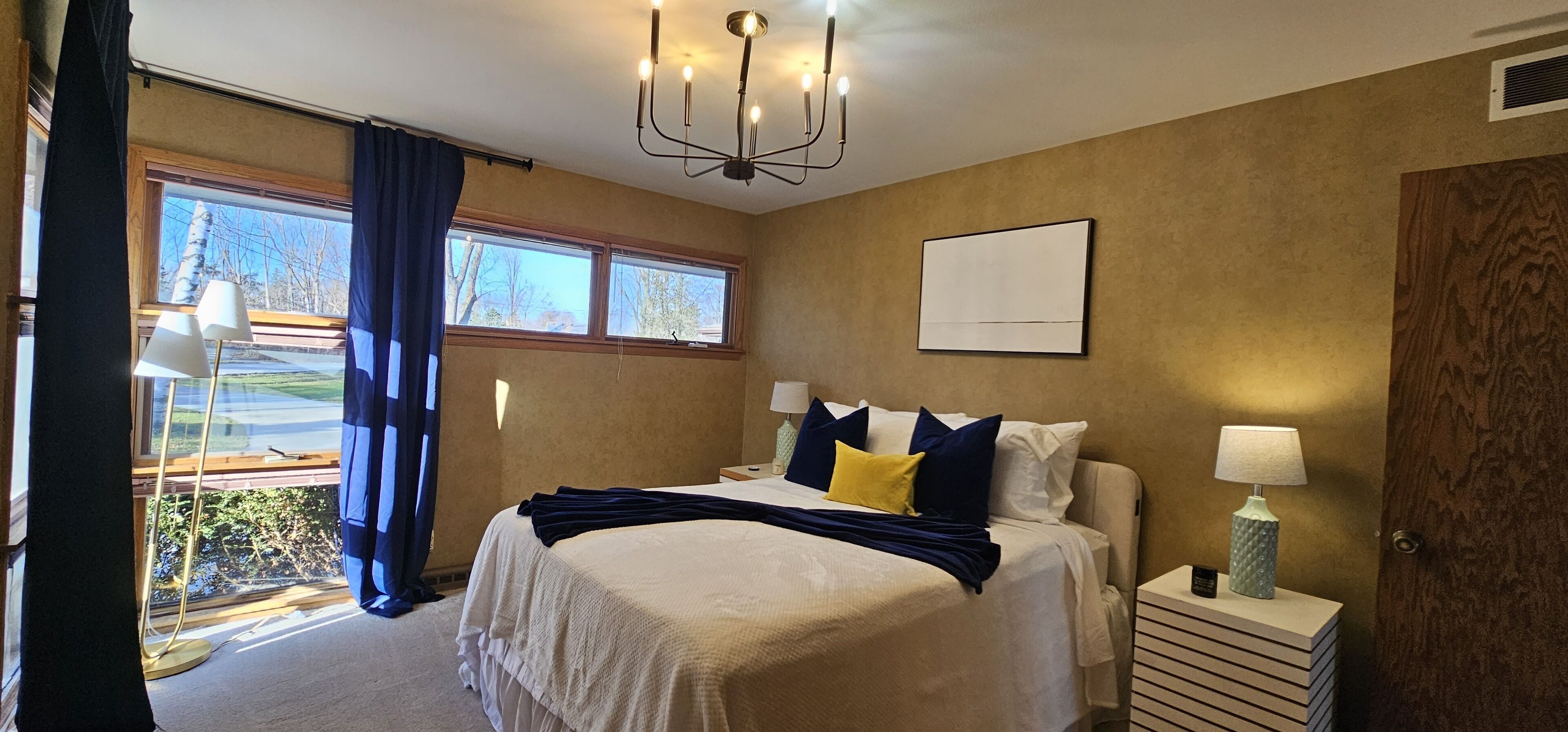 Ein Schlafzimmer mit einem großen Bett, einer Chandelüster, einem Nachttisch mit Lampe und einem Fenster mit Vorhängen.