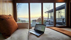 A laptop on a cushioned bench by a large window with a view of a building and a body of water.