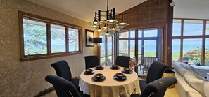A dining area with a round table set for six, large windows offering a view of the sea, and a cozy seating area with a sofa and armchairs.