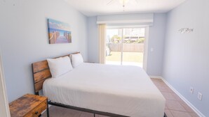 Een slaapkamer met een bed, nachtkastje, plafondventilator, schilderij en een schuifdeur naar buiten.