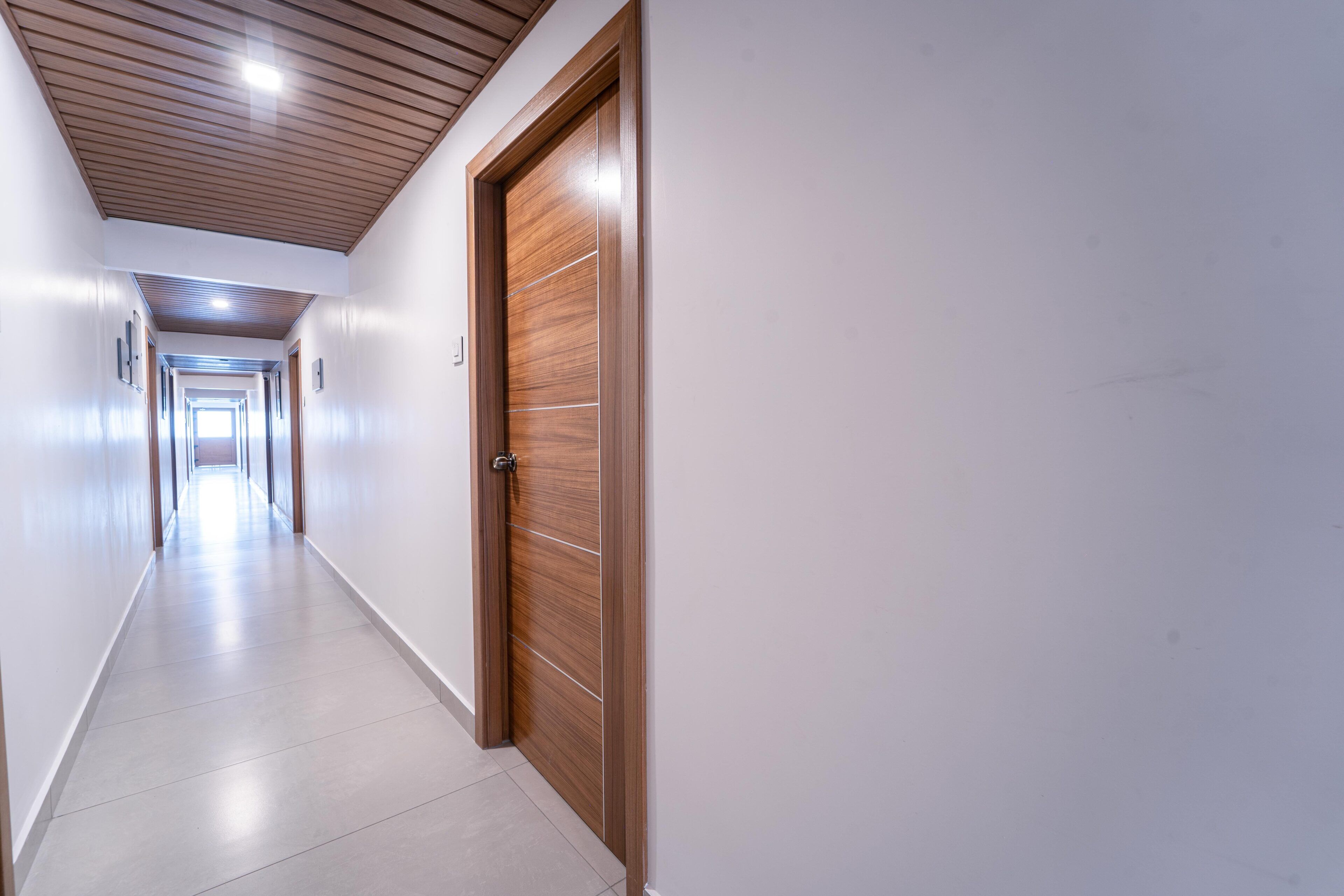 Un pasillo largo y estrecho con una puerta de madera a la derecha, paredes blancas y piso de baldosas.